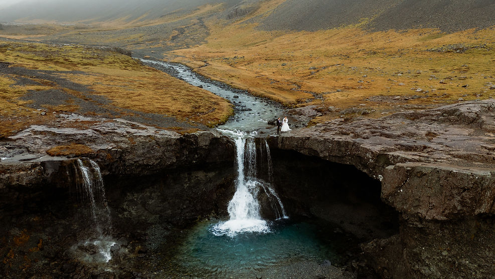 During Iceland Elopement, bride and groom stand near a waterfall on rocky terrain, surrounded by a mountainous, misty landscape with autumn colors. Romantic scene.