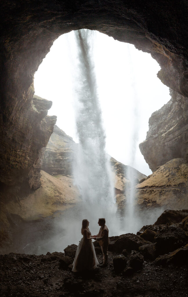 A couple stands hand in hand in a cave behind a cascading waterfall. Silhouetted figures, with rocky terrain and misty atmosphere. Romantic mood for the best place to elope in iceland.