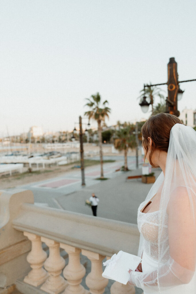 Bride in white dress and veil stands on balcony in Barcelona, holding a notebook. She looks at a person on the promenade with palm trees and boats.