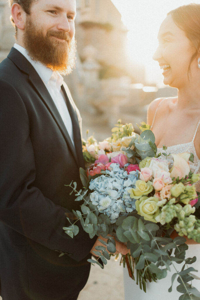 Bride and groom smiling, holding vibrant bouquet, against sunset backdrop. Bride in sparkling dress, groom in black suit, joyful mood.