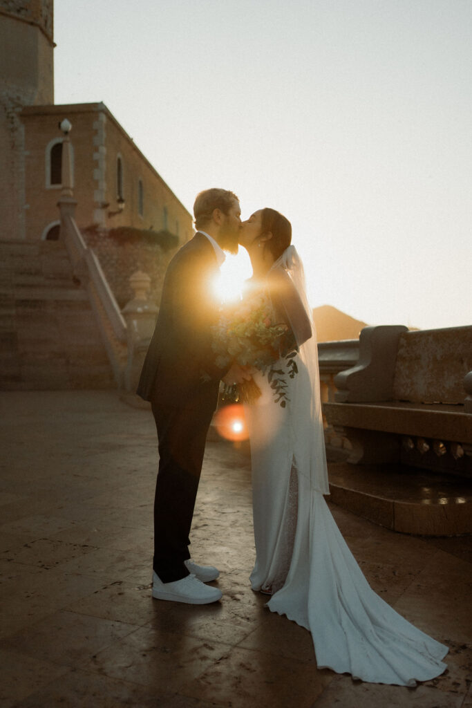 A couple kisses at sunset in Barcelona, silhouetted by sunlight. They are dressed in wedding attire, standing on stone steps with a historic building behind them.