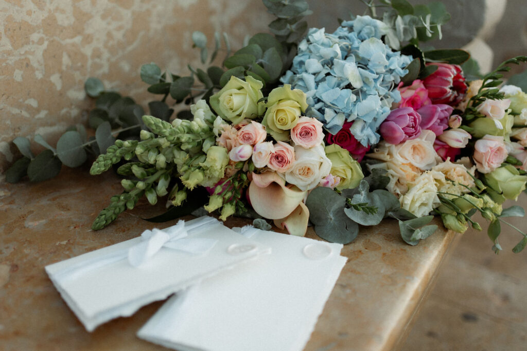 Bright elopement bouquet of roses and hydrangeas on a stone bench with white envelopes. Soft colors create a serene, romantic mood.