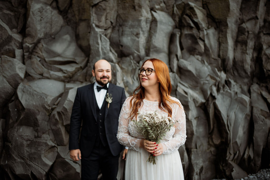 Bride in white lace dress holds elopement flowers, standing with a smiling man in a black suit. They are in front of a rocky wall in iceland.