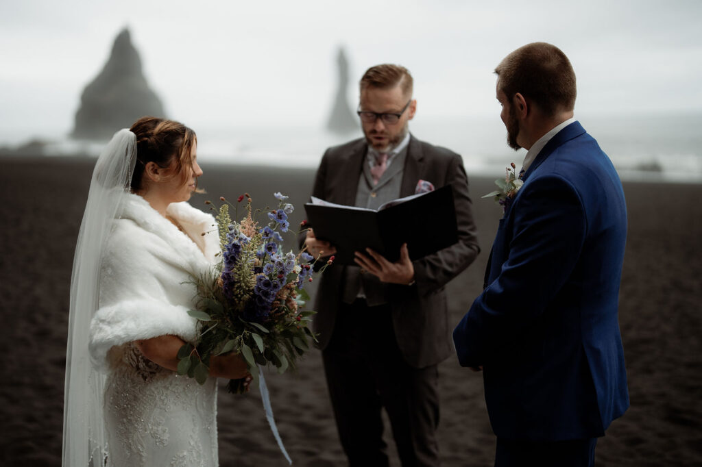 Bride and groom stand on a black sand beach as officiant reads. The bride holds a bouquet of purple flowers. 