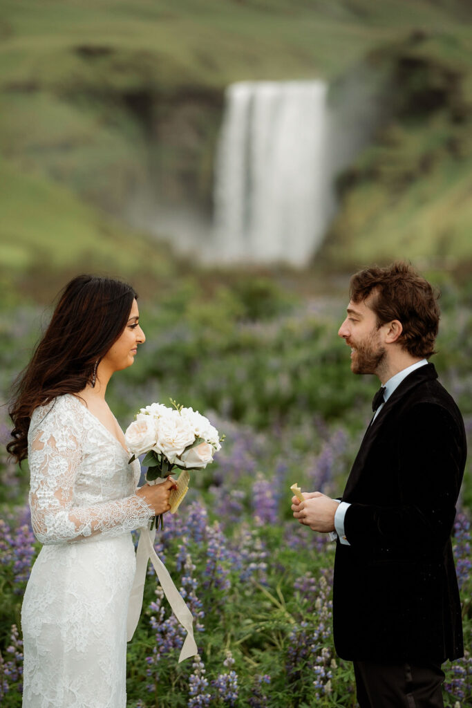 Bride in white lace dress holding elopement flowers faces groom in black suit, set in a lush field with a waterfall in the background. Romantic mood.