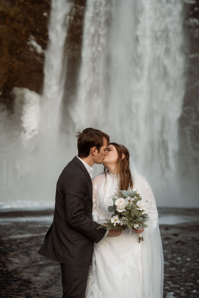 bride and groom kiss while holding elopement bouquet in front of waterfall