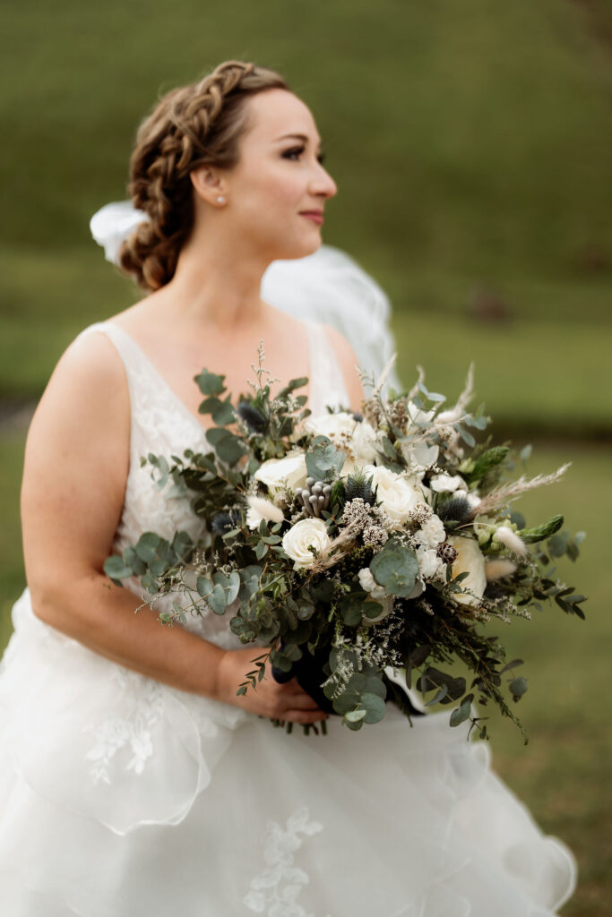 Bride with braided hair holds an elopement bouquet of green and white flowers, wearing a lace dress. She stands outdoors, looking serene against a grassy backdrop.