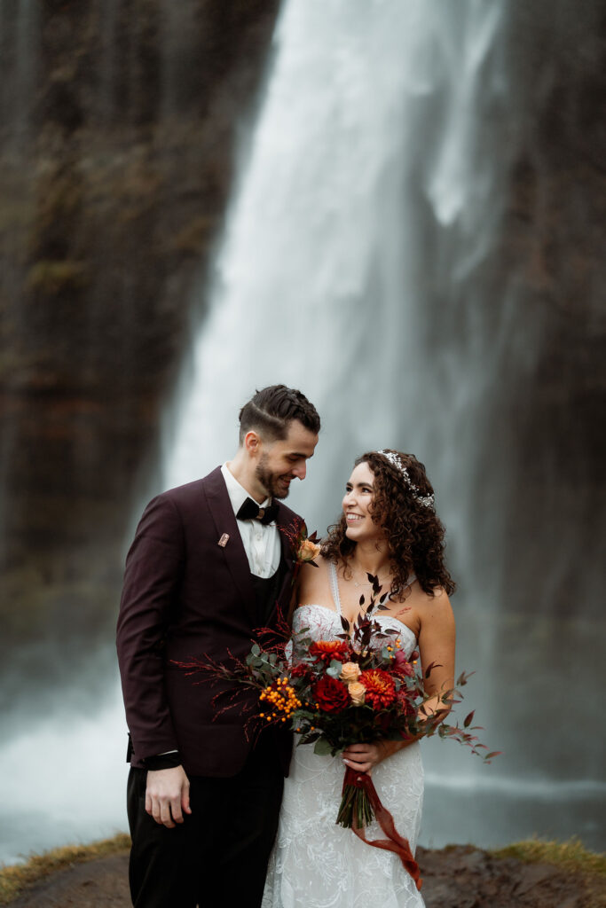Bride and groom smiling by a waterfall. Bride holds a vibrant bouquet. Groom in a dark suit. 