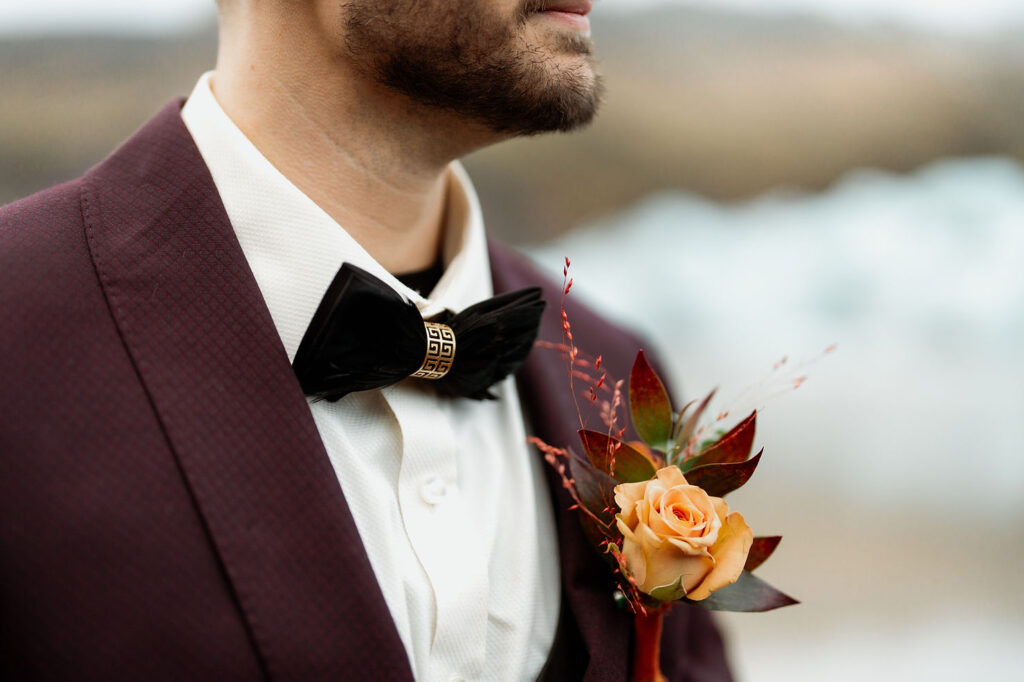 Close-up of a person in a maroon suit and white shirt, wearing a black bow tie with a gold detail, and an orange rose boutonniere.