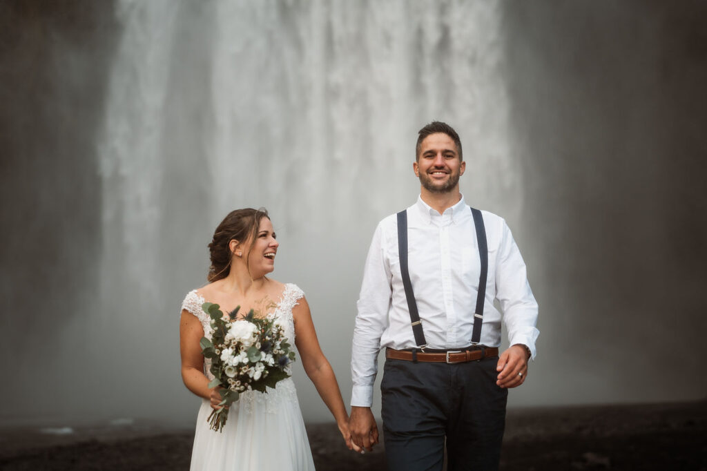 bride and groom hold hands and smile as they walk in front of waterfall during iceland elopement