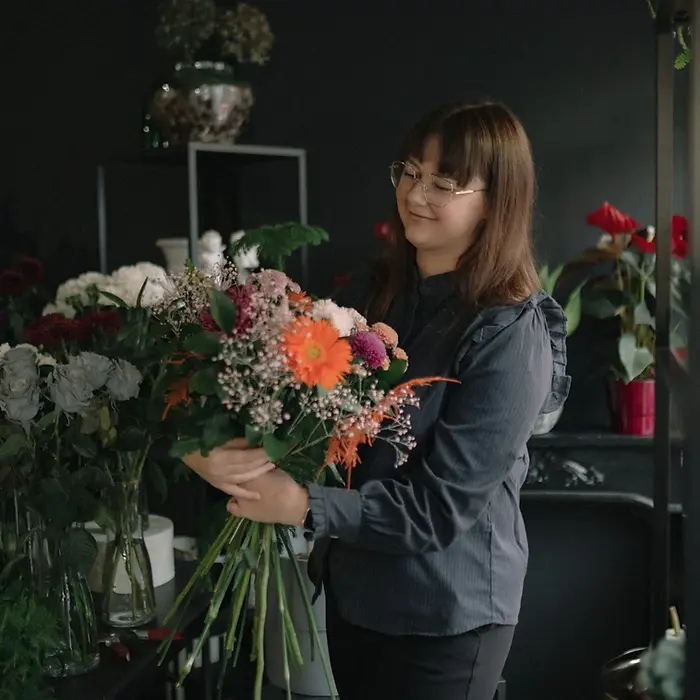 An iceland florist makes a vibrant elopement bouquet of mixed flowers, smiling in a dark floral shop. The background features various plants and muted colors.