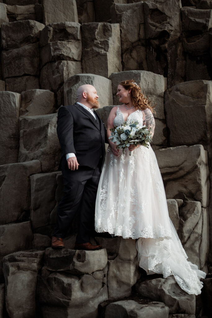 Bride in a white lace dress and groom in a black suit stand on rocky terrain, gazing at each other, holding an elopement bouquet, with a joyful mood.