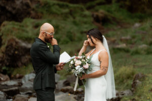 A bride and groom exchange emotional vows outdoors, surrounded by greenery. She holds an elopement bouquet, both wiping tears, set in a serene grassy area.
