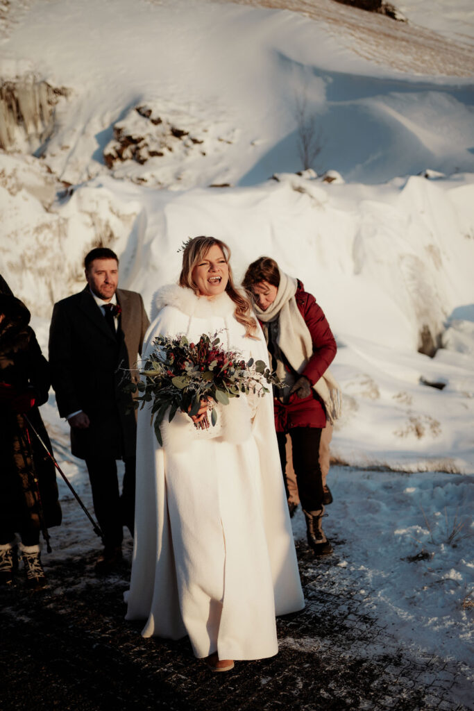 Bride in a white cloak holding an elopement bouquet, smiling in a snowy landscape. Two people stand behind her, one in a red coat. Sunny winter day.