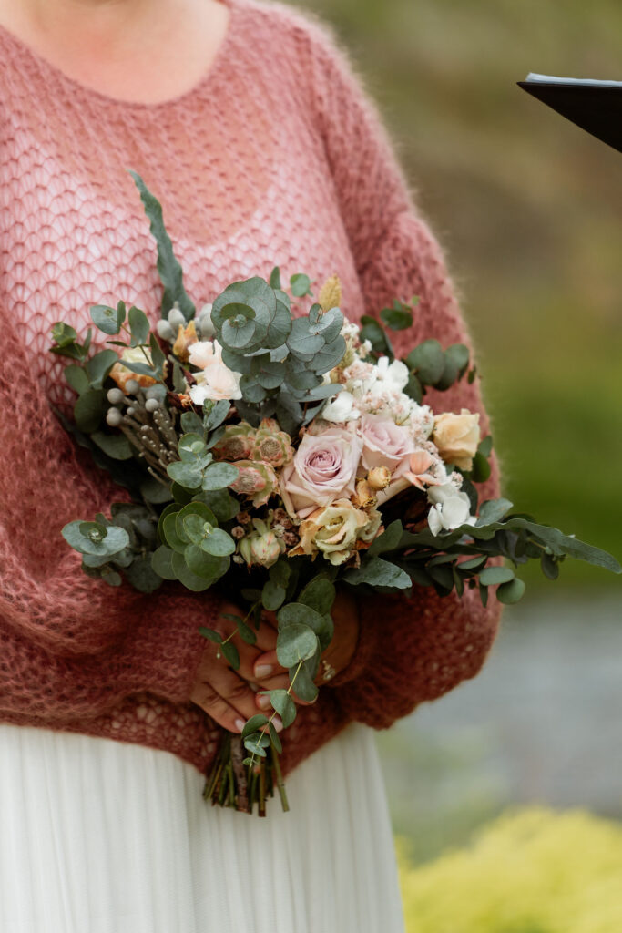 Person in pink sweater holding iceland elopement bouquet with pastel roses and greenery, outdoors. 