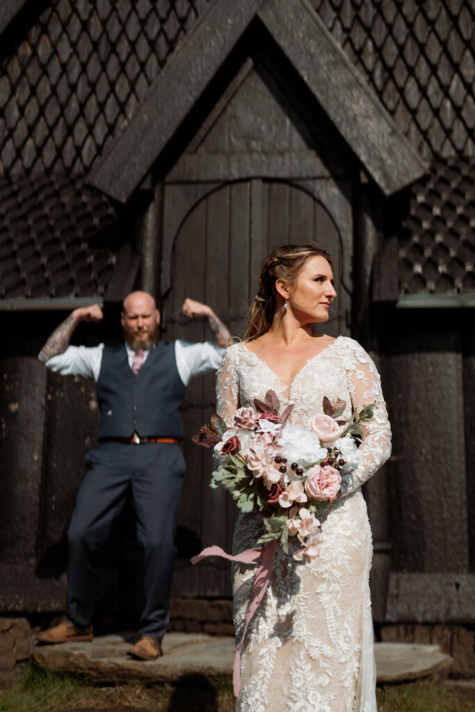 Bride in lace dress holds elopement bouquet, stands in front of wooden building. Man in vest flexes in background, sunny day, relaxed mood.
