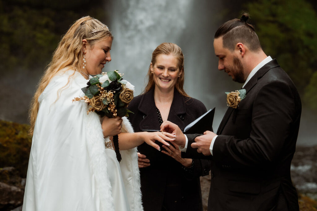 Bride in lace dress holds elopement bouquet, stands in front of wooden building. Man in vest flexes in background, sunny day, relaxed mood.
