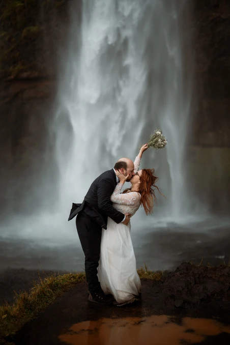 bride and groom stand at the bottom of a waterfall while bride holds up elopement bouquet