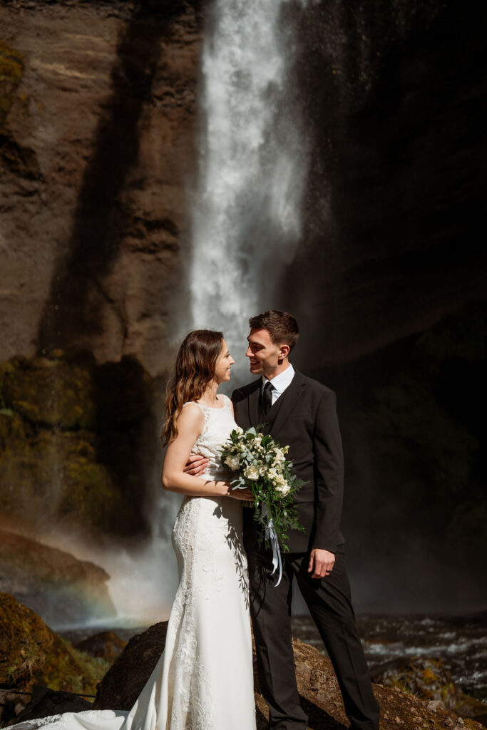 Bride and groom embracing in front of a waterfall, she holds an iceland wedding bouquet. Sunlit scene with a hint of a rainbow in the background.