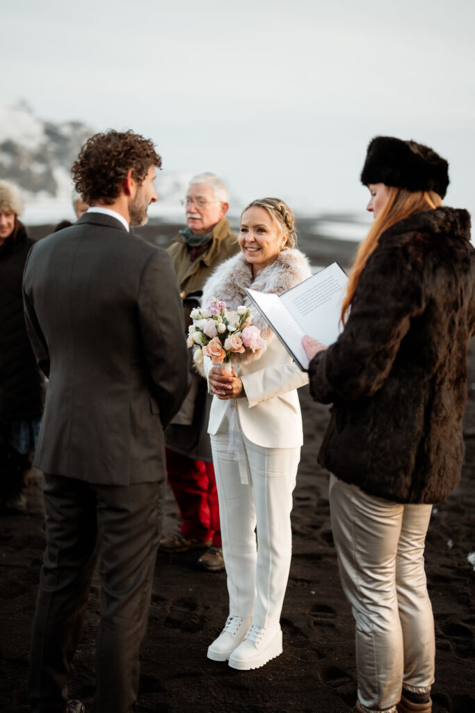 Bride in white suit holding elopement flowers, smiling at groom in suit on a black sand beach during a ceremony. An officiant reads from a book.