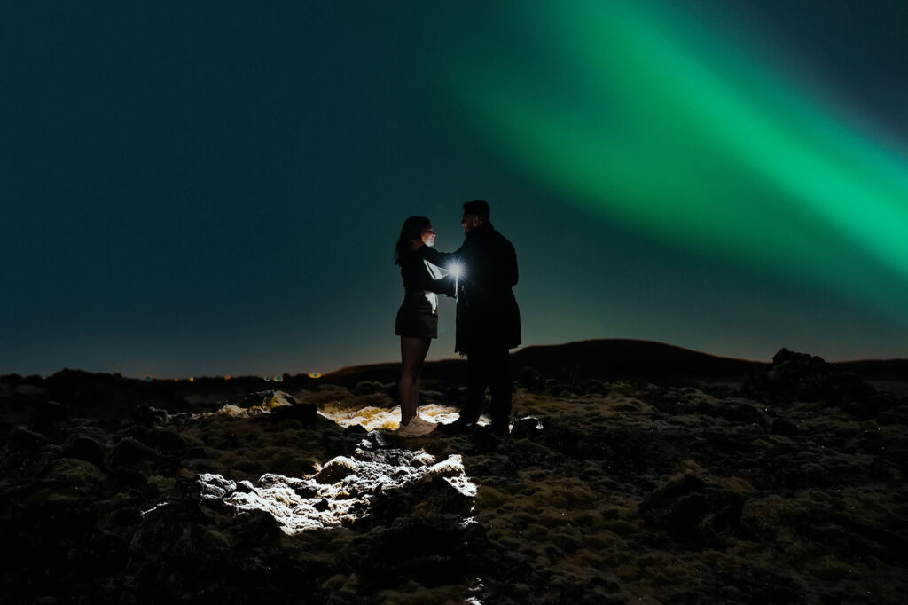 Two people stand under a vibrant green aurora in a dark, rocky landscape, illuminated by a flashlight during northern lights proposal in iceland