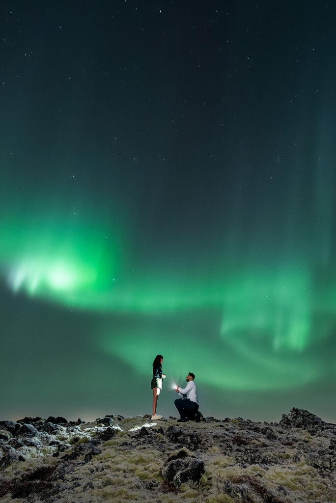 Man kneels proposing to woman under green aurora borealis in iceland landscape. Starry night sky shows romantic and magical ambiance.