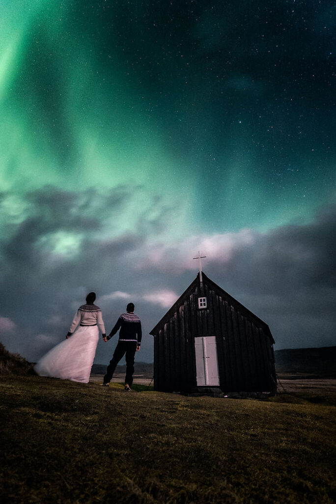 bride and groom holding hands by a small black chapel under vibrant green northern lights, grassy field, nighttime; peaceful in iceland.