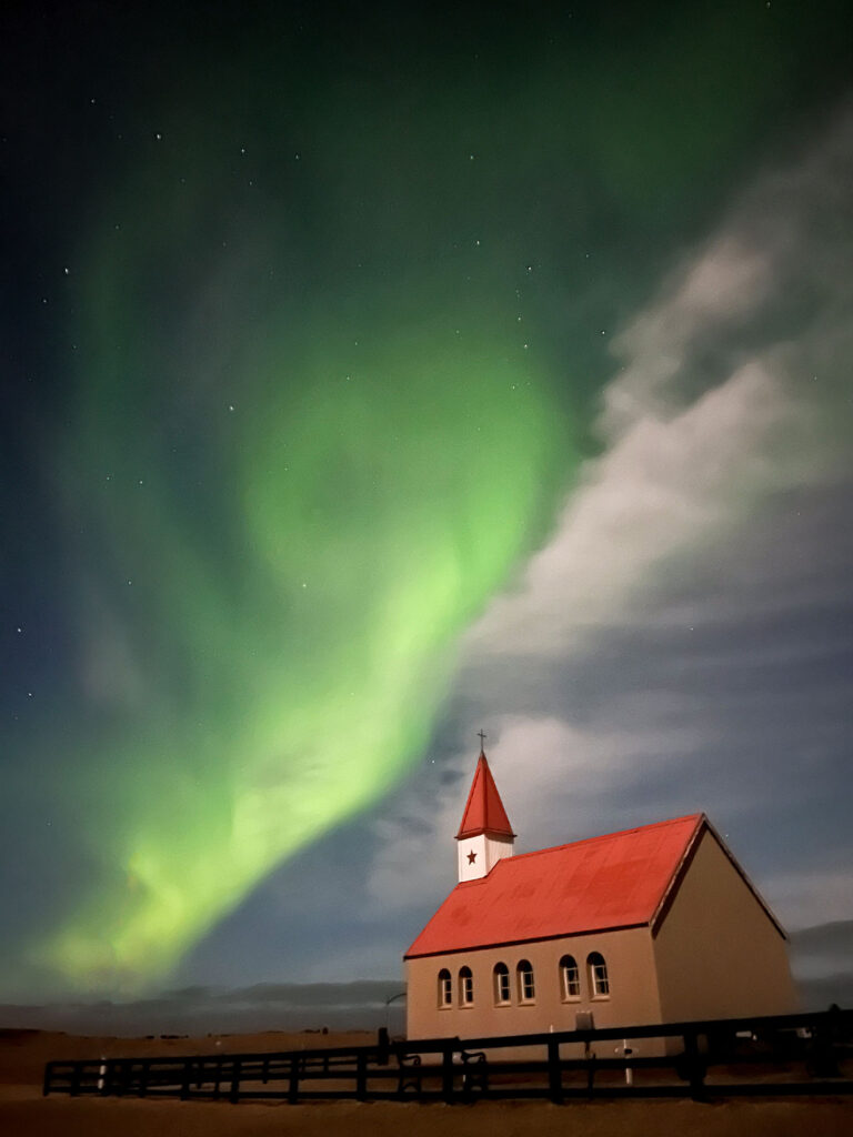 A church with a red roof under vibrant green Northern Lights in a night sky, fenced foreground. Serene and awe-inspiring atmosphere.