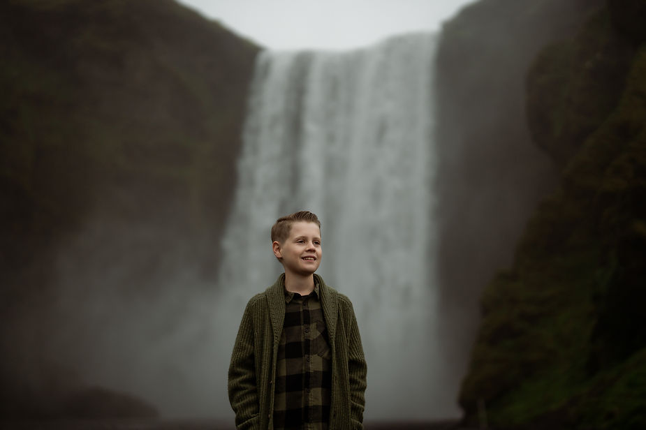 Young boy stands in front of waterfall during family photos by Iceland wedding photographer.