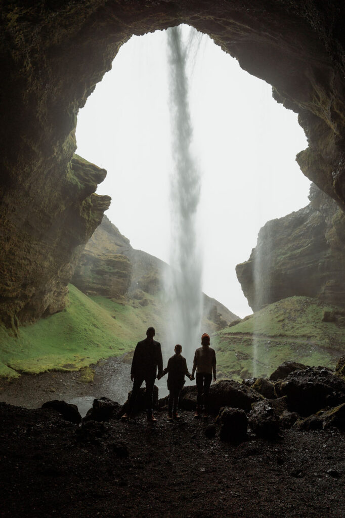 family of 3 stand in the shadows, behind a waterfall, during Iceland Family Photos.
