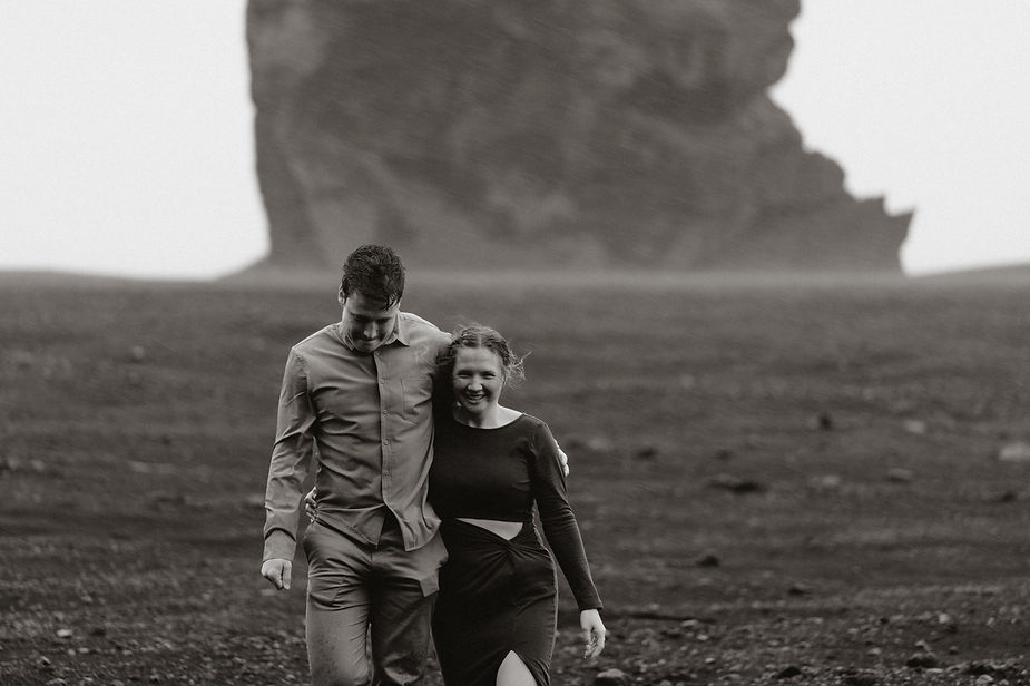 engaged couple walks along black sand with giant rock in the background during iceland engagement photo shoot.
