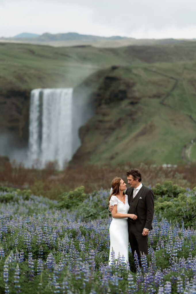 married couple stands in field of flowers and waterfall during iceland wedding photo shoot.
