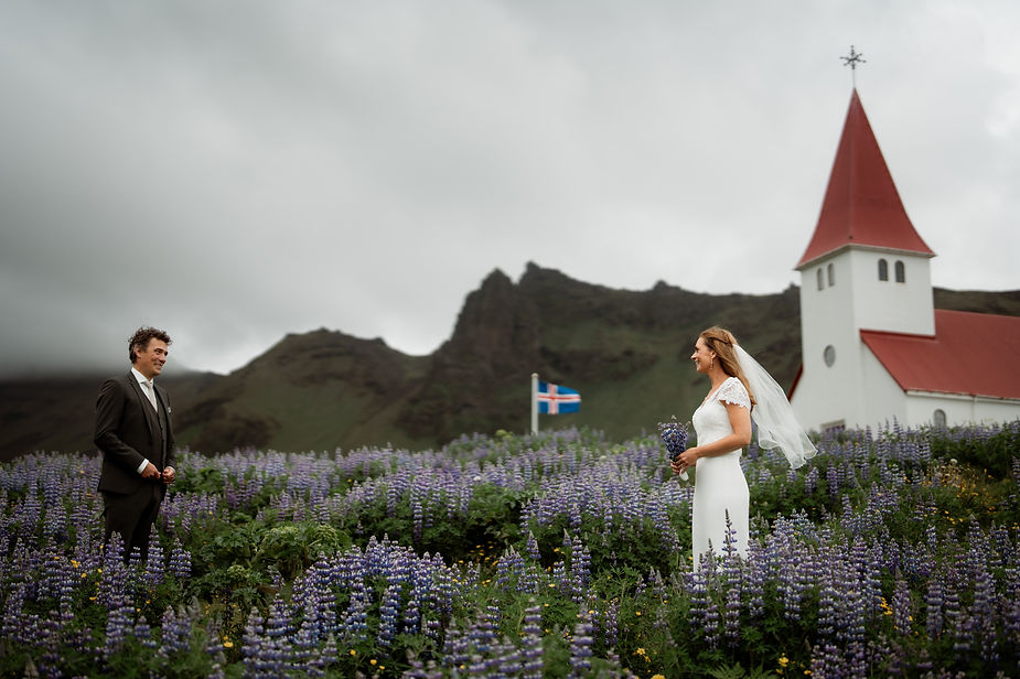 bride and groom stand in field of purple flowers in front of church during iceland wedding.