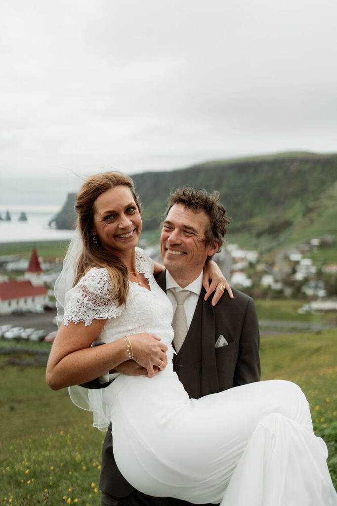 groom holds bride with town in the background, during iceland wedding photos.