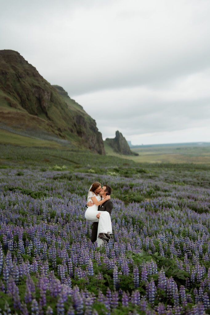groom holds bride in field of purple flowers on iceland coast during adventure iceland wedding.