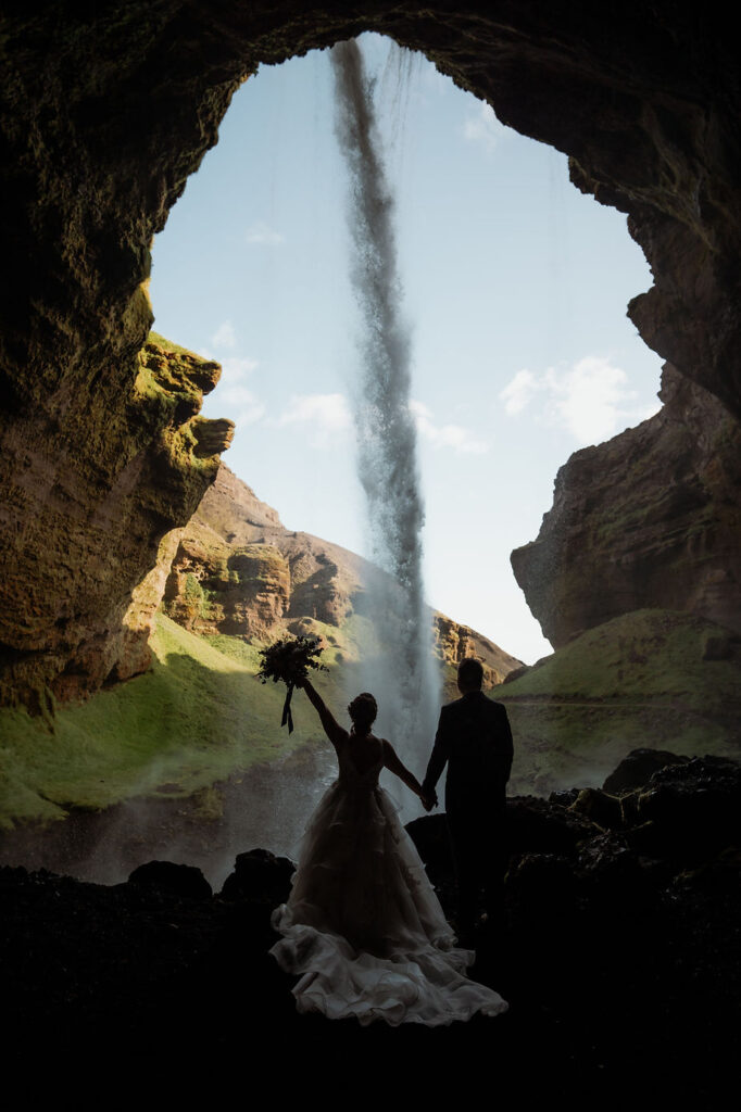 Newlywed couple stands behind waterfall during adventure wedding in iceland.