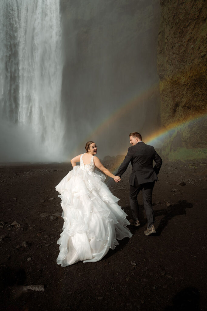 newlywed couple runs towards waterfall with rainbow in the background, during iceland adventure wedding.
