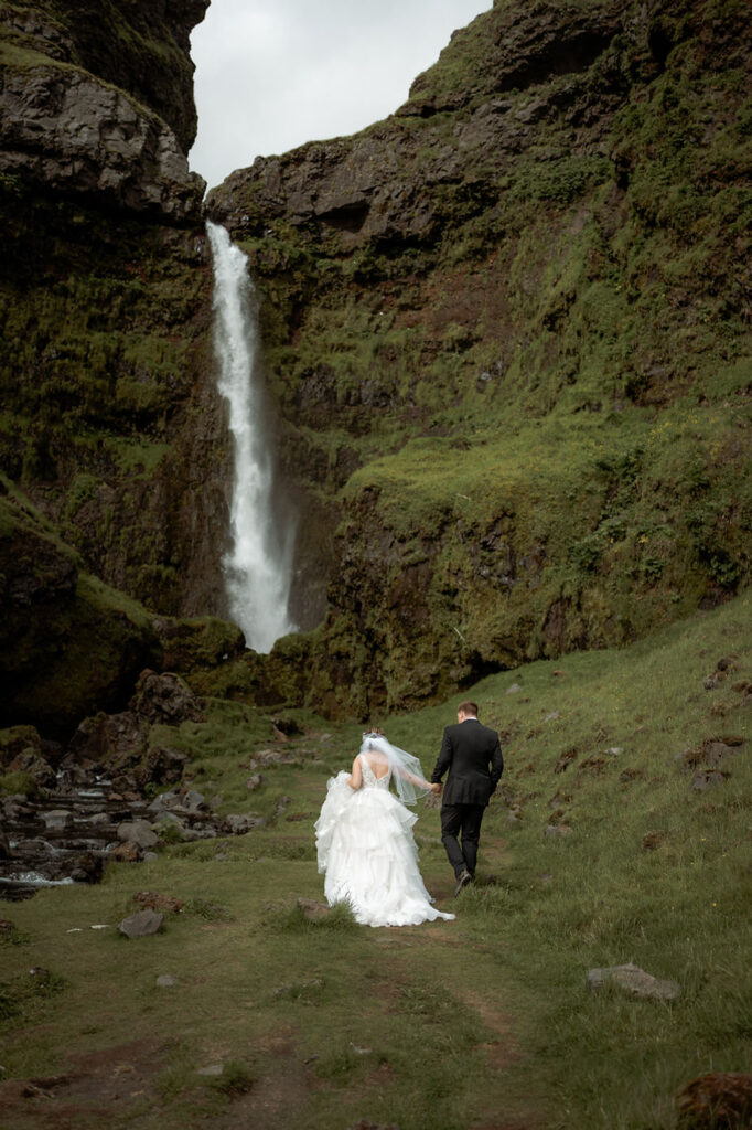 Bride and groom walk towards waterfall during iceland elopement photos.