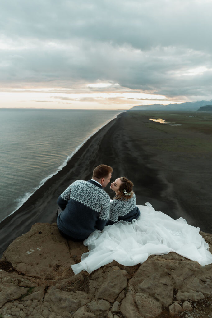 bride and groom sit, in sweaters, on a cliff edge, overlooking the black sand beach during adventure elopement in iceland.