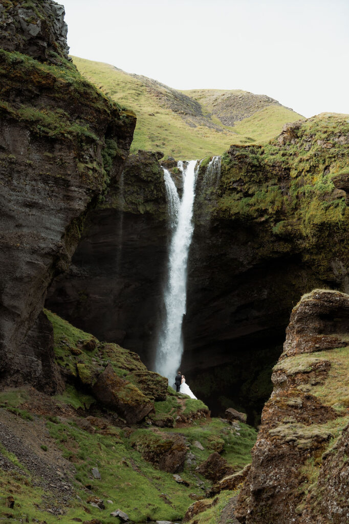 bride and groom stand at the base of waterfall during iceland adventure elopement.