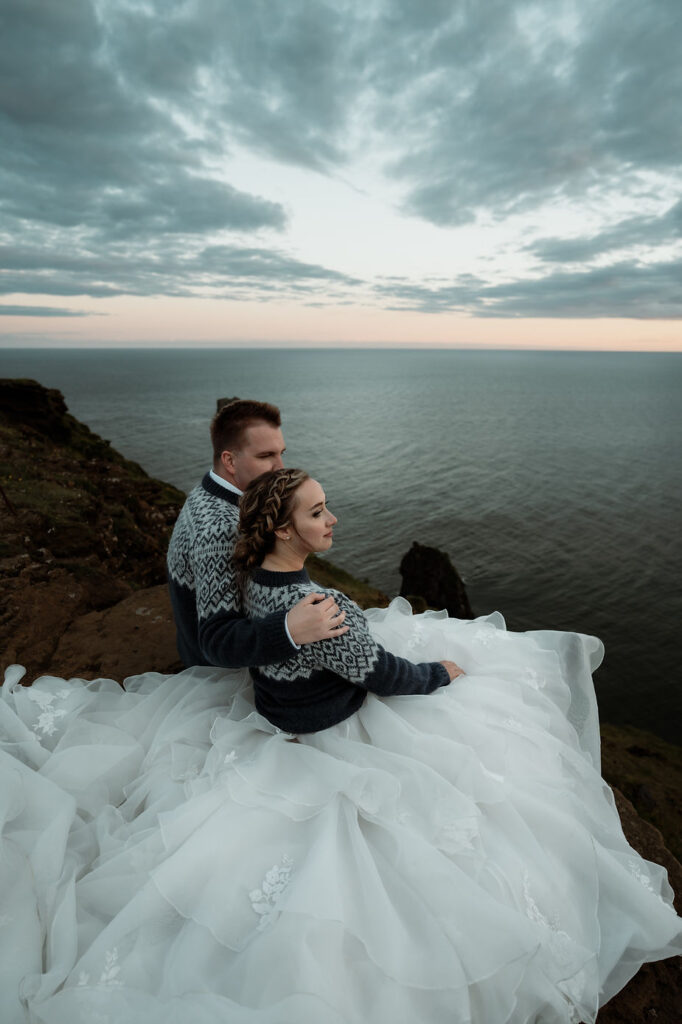 newlywed couple sits on cliff edge and watches the sunset during dreamy iceland elopement.