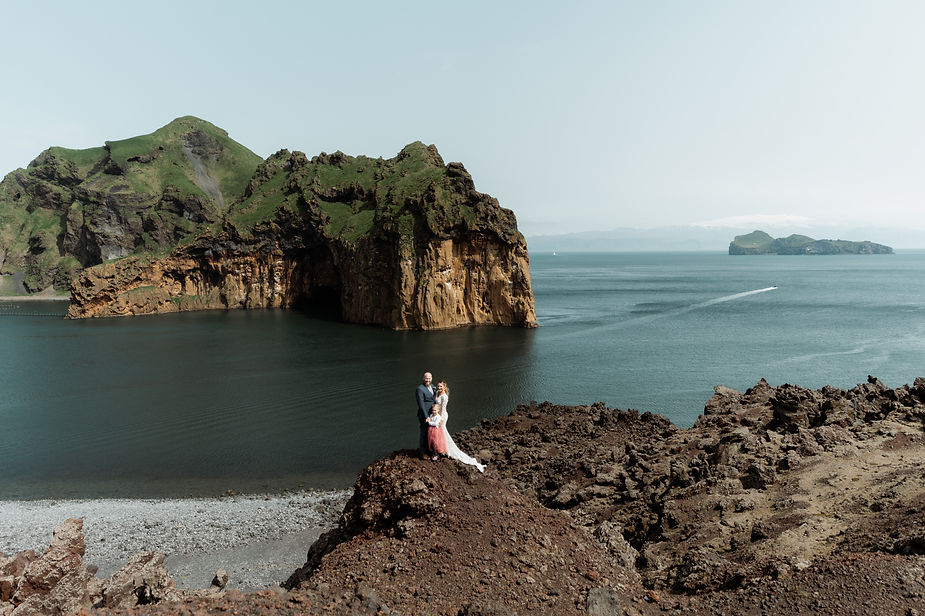 Bride, groom, and daughter stand on rocky ledge overlooking the westman islands during elopement.