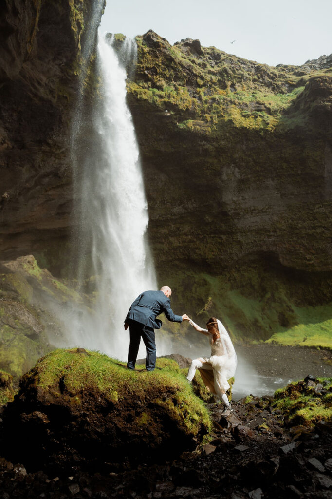 Groom helps bride up rock in front of waterfall during iceland elopement.