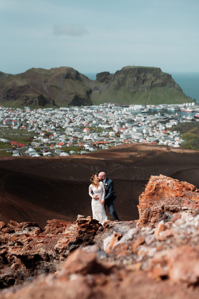 Bride and groom stand on vibrant orange cliff edge with westman islands in the background during iceland wedding