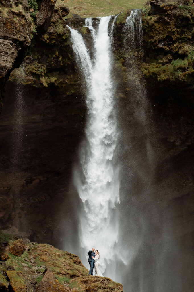 bride and groom stand at the bottom of waterfall during iceland adventure wedding photos.