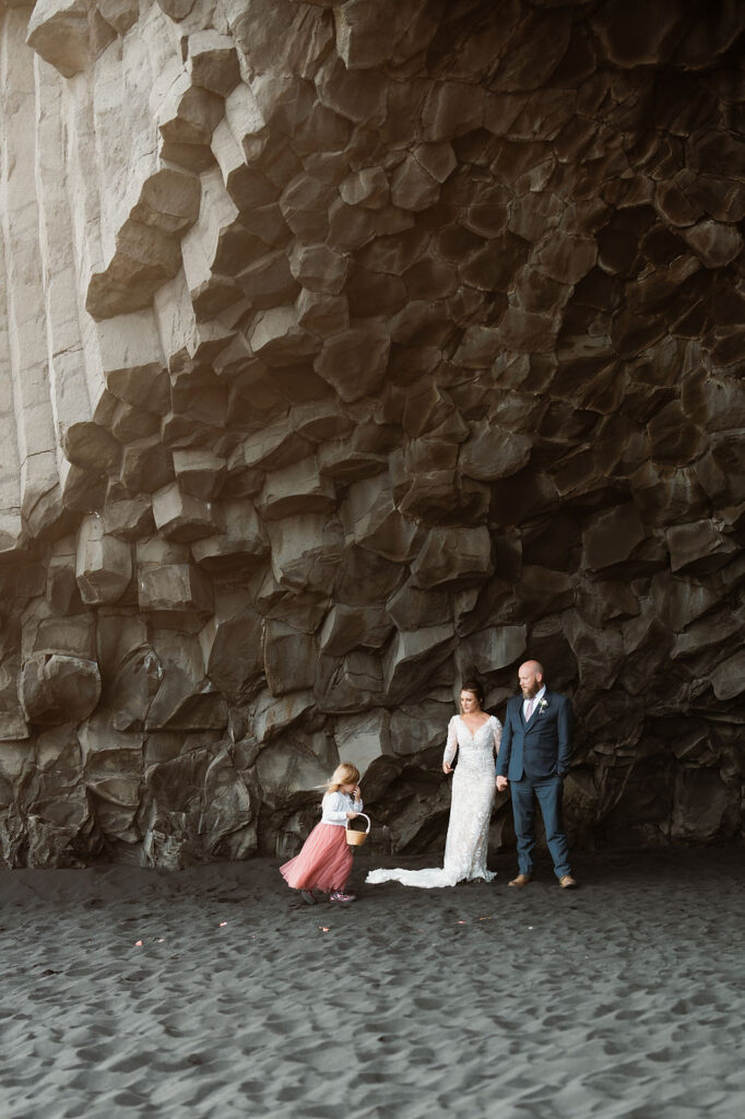 bride, groom, and daughter stand under rock formation during iceland wedding photo shoot.