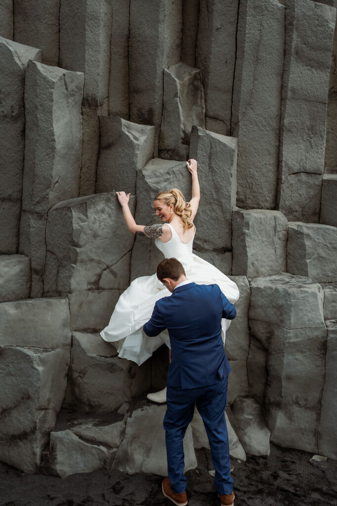 groom helps bride climb up rocks during iceland wedding adventure.
