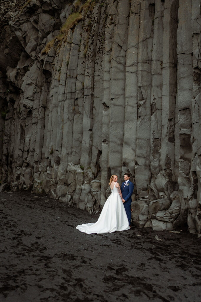 bride and groom stand under rock formations during iceland wedding day adventure.