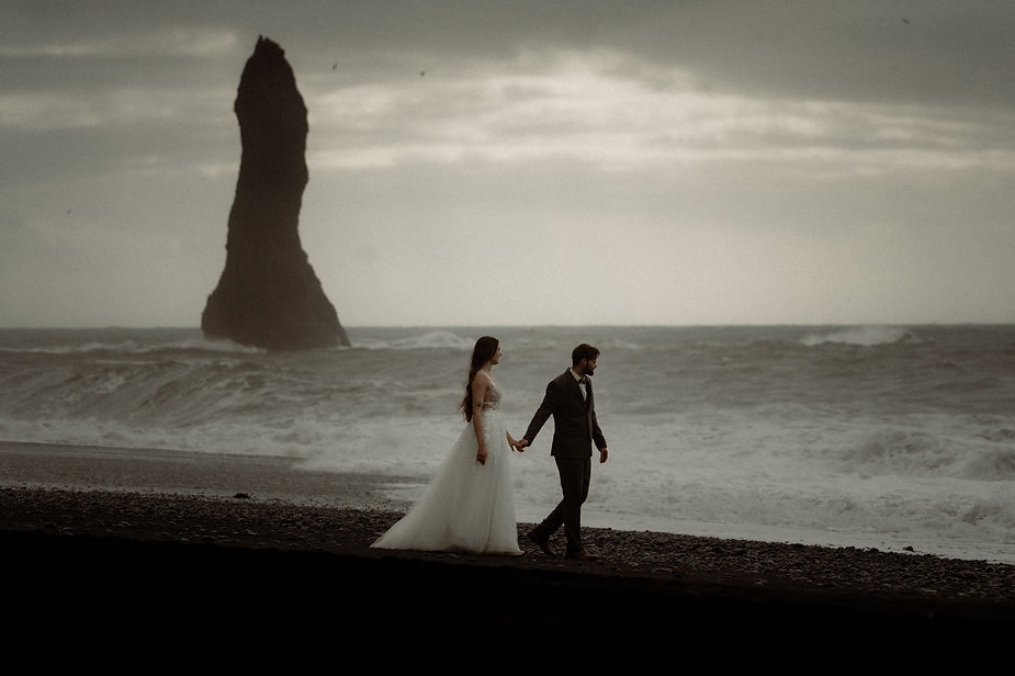 bride and groom walk along black sand beach in iceland during honeymoon.