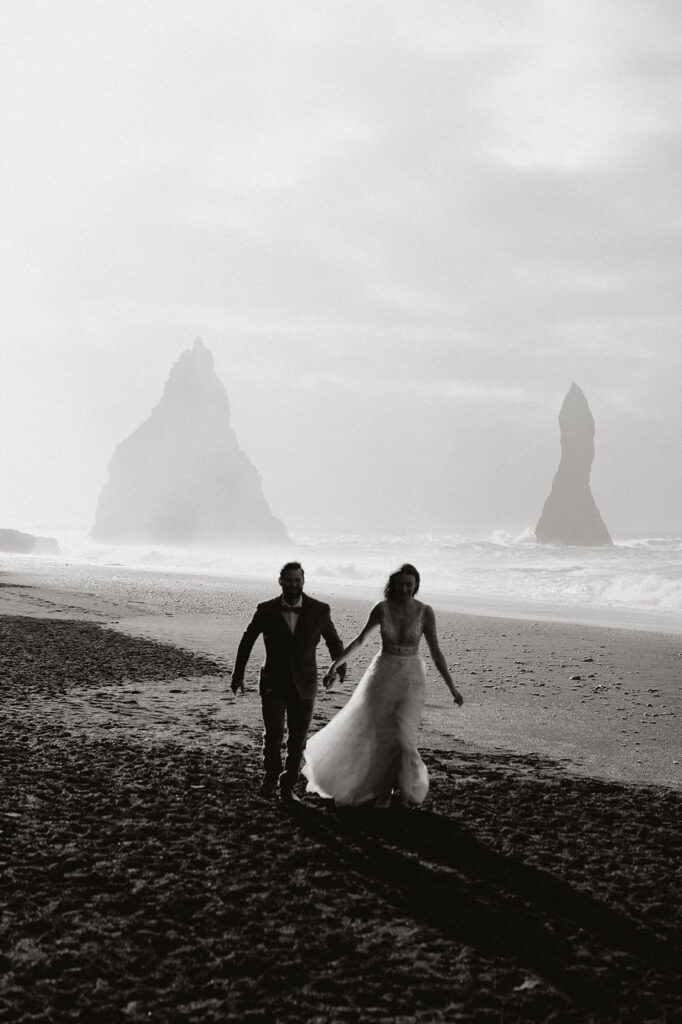 bride and groom run along black sand beach during iceland honeymoon.